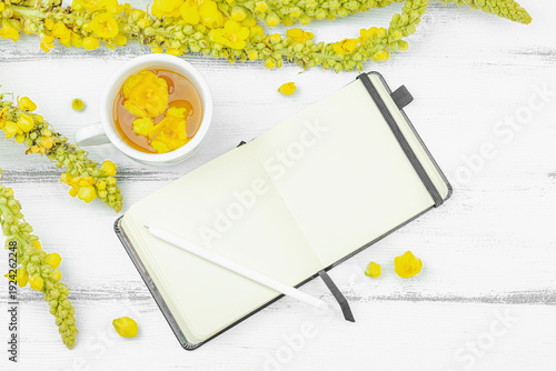 Cup of herbal tea with mullein flowers and blank notebook with pen on white wooden table