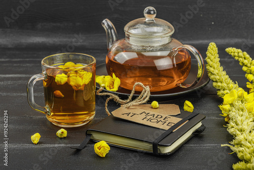 Glass cup of mullein flower tea with teapot and notebook on black wooden table, healthy drink