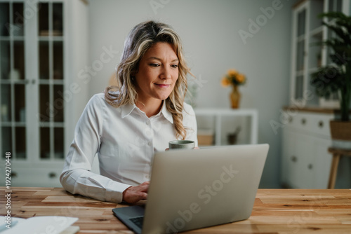 Wallpaper Mural Mature woman smiling, holding coffee while working on laptop at home, remote business professional Torontodigital.ca