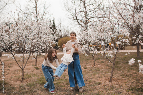 Happy mother with two daughters wearing denim clothes having fun in blooming almond trees garden outdoors. Motherhood. Looking at camera.