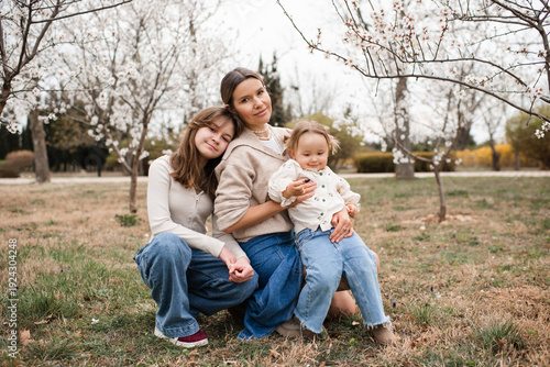 Happy mother with two daughters wearing denim pants in blooming garden outdoors. Motherhood. Looking at camera.