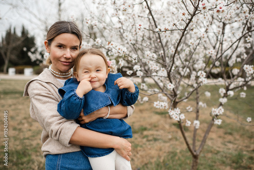 Stylish woman holding baby girl 1-2 year old wearing denim dress having fun over blooming almond trees with flowers outdoors. Motherhood. Springtime.