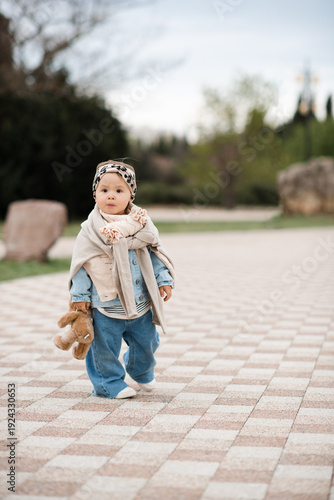Stylish baby girl 1-2 year old wearing trendy clothes with denim pants and headband holding teddy bear toy in park outdoors. Springtime. Childhood.