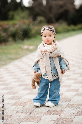 Stylish baby girl 1-2 year old wearing trendy clothes with denim pants and headband holding teddy bear toy in park outdoors. Springtime. Childhood.