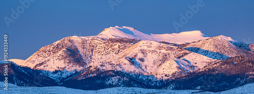 Mt. Rose, located near Reno, Nevada, is depicted covered in fresh snow at dawn. The mountain's rugged peaks are illuminated with a soft pink-orange glow from the early morning light.