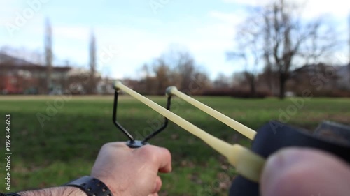 Slow motion close up of hands pulling back a slingshot and releasing a small stone outdoors. Elastic band stretches tightly before the projectile shoots forward in dramatic detail.