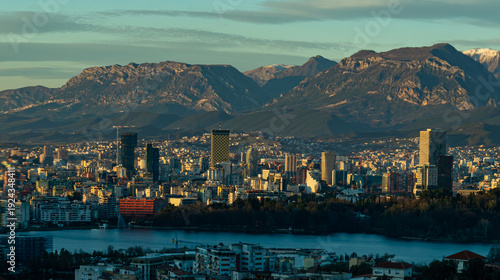 Wide-angle panoramic view of the Tirana city skyline at sunset, featuring the Artificial Lake in the foreground and the snow-capped Dajti Mountains under a soft evening sky.