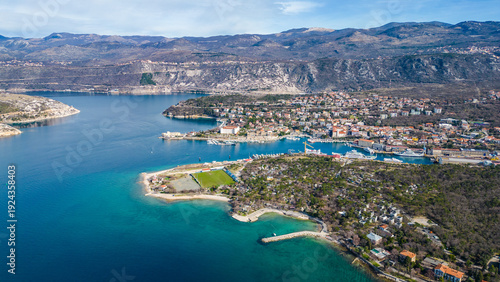 An aerial view of a vibrant Croatian coastal peninsula, featuring turquoise waters, a football field, and a bustling camp, bathed in bright morning sun. Kraljevica