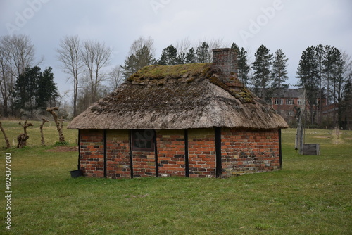 Side view of old brick house with thatched roof and chimney, standing in green grass field, grey skies, autumn time, Lyngby, near Copenhagen, Denmark