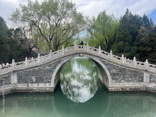 Side horizontal view of Chinese stone arch bridge, bridge over green water, trees in back, white clouds in sky, Beijing Summer Palace, Beijing, China 