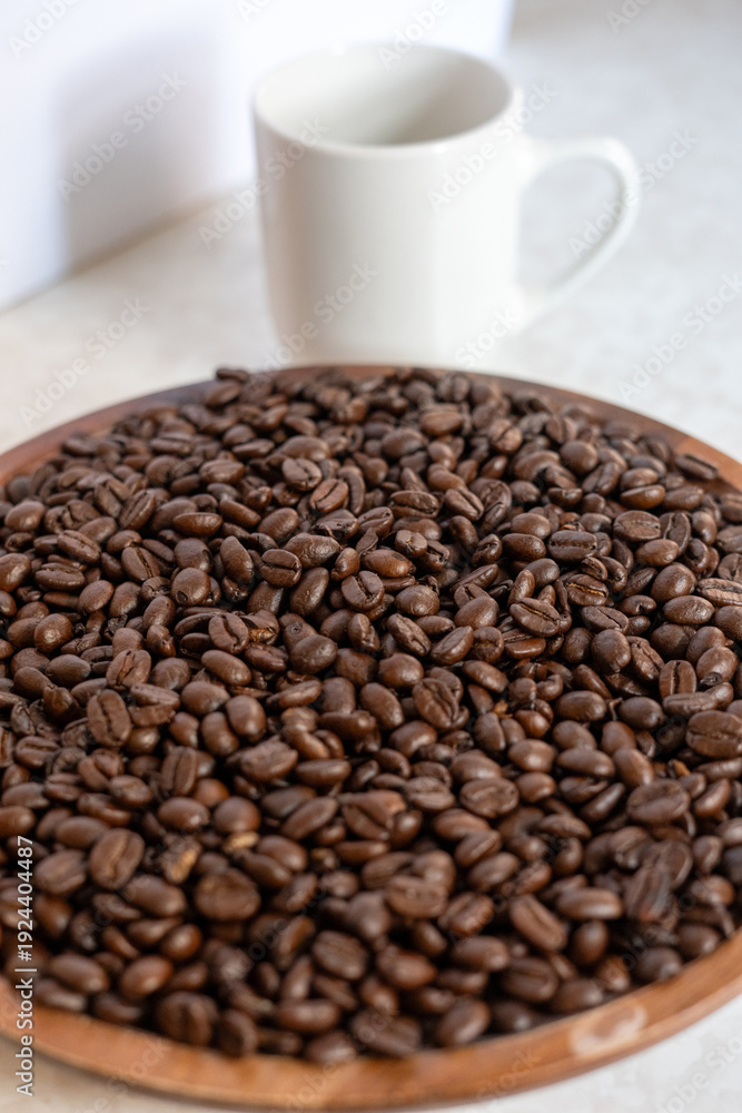 Fototapeta premium Roasted coffee beans in a wooden tray with a white ceramic cup on a light marble table.