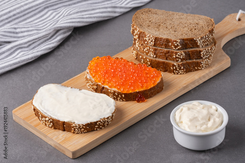 Slices of brown bread with spreads of cream and red caviar on a wooden board in a kitchen setting with a striped cloth