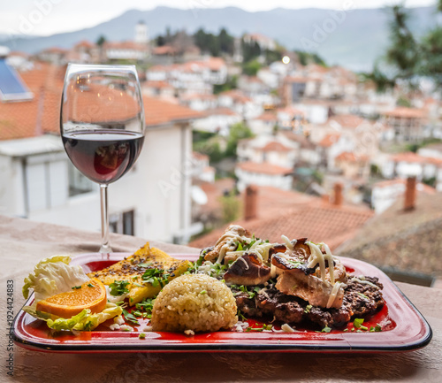 Photos A delicious Balkan dinner platter with grilled meat, rice, polenta, and salad, paired with a glass of red wine, all overlooking a blurred Ohrid city panorama