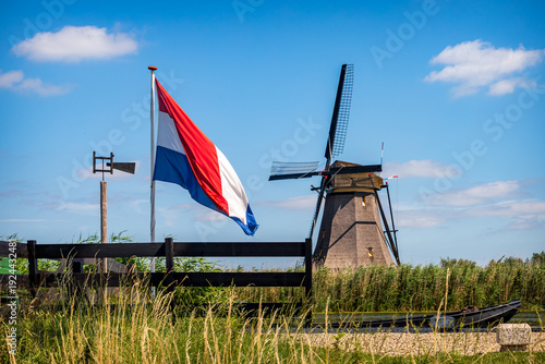 Dutch flag and traditional windmill at Kinderdijk in the Netherlands with reeds and canal, iconic UNESCO World Heritage polder scene on a sunny day