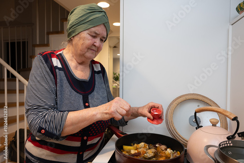 Senior woman preparing a family dinner in her home kitchen, adding salt to a hot stew on the stove. Mother's Day concept