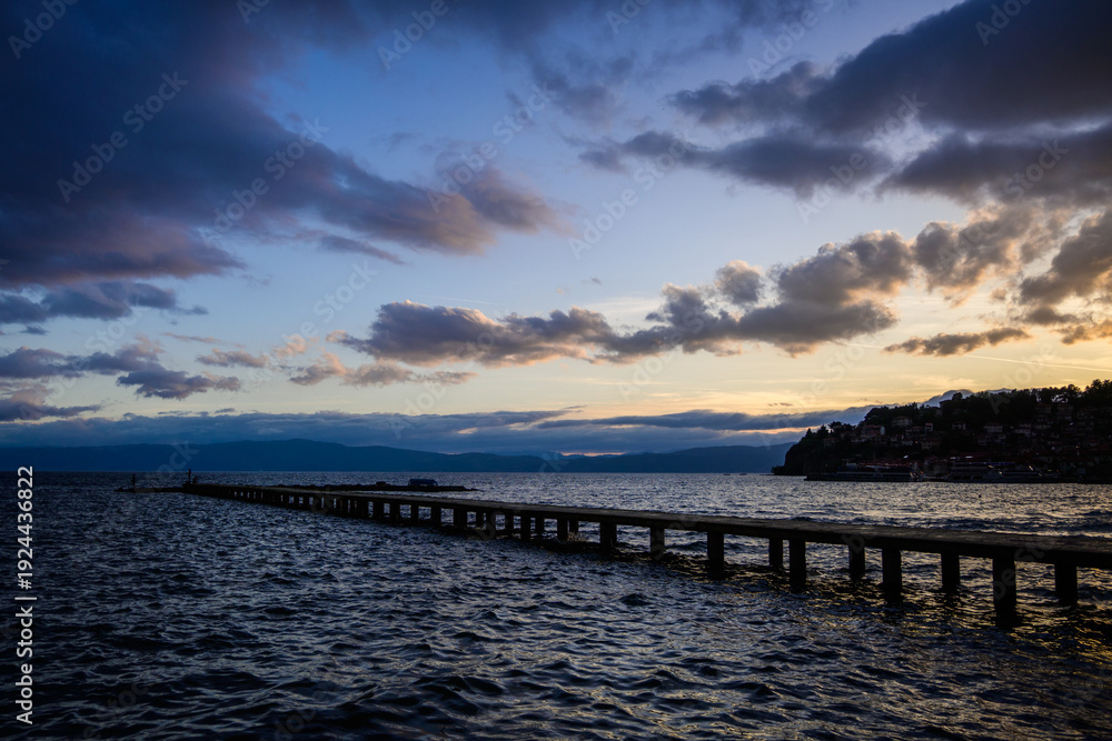 Obraz premium A long pier extends into the choppy, dark waters of Lake Ohrid under a dramatic twilight sky, filled with striking cloud formations and distant town lights.