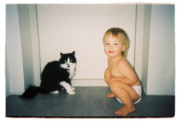 Toddler sitting on floor next to black and white cat, analog film