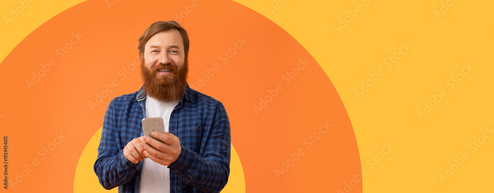 Obraz premium A man stands in front of a bright orange background. He holds a smartphone in his hands and smiles. The scene is simple and eye-catching.