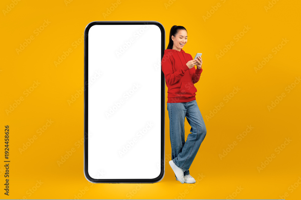 © Prostock-studio - A young woman stands next to a large blank phone display. She is looking at her smartphone and smiling. The background is bright yellow, creating a lively atmosphere.