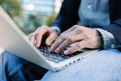 Close-up of adult man typing on laptop outdoors. Focused professional working digitally in an urban park. Business and technology concept.