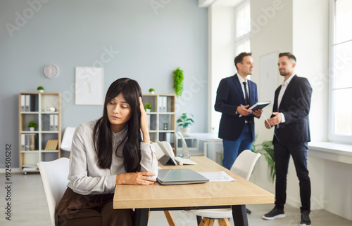 Slika na platnu An employee sits apart, showing isolation and stress as colleagues chat, hinting at gossip, exclusion, burnout, loneliness, and harassment in a workplace
