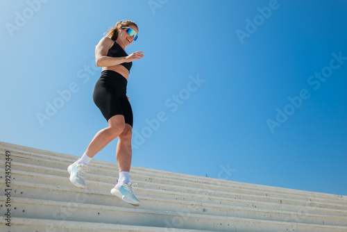Woman exercises on stairs under clear blue sky during daytime while wearing sports gear and sunglasses