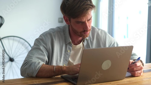 Side view handsome young businessman working with computer remotely, sitting at wooden table in office. Pleasant happy man communicating in social network, searching information online.

