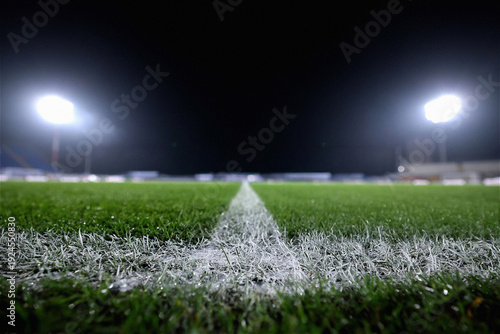 Close-up low-angle view of a white painted line on a green grass sports field at night under bright stadium floodlights. This atmospheric shot captures the essence of competition, professional sports,