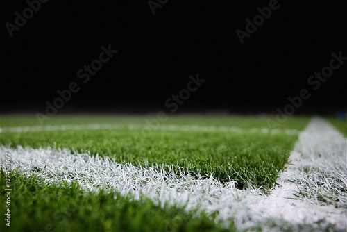 A professional close-up of a green sports field at night, featuring crisp white painted boundary lines on lush turf. The low-angle shot captures the texture of the grass under stadium lighting, with a