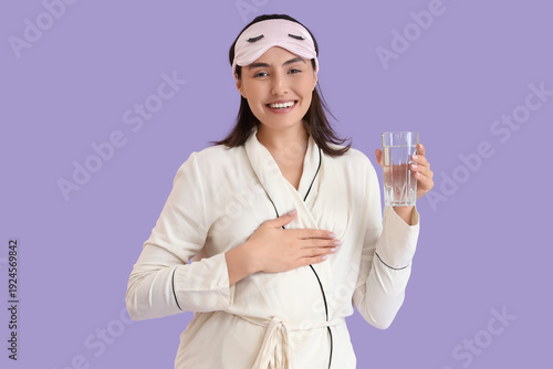 Young woman in pajamas with sleeping mask and glass of water on lilac background