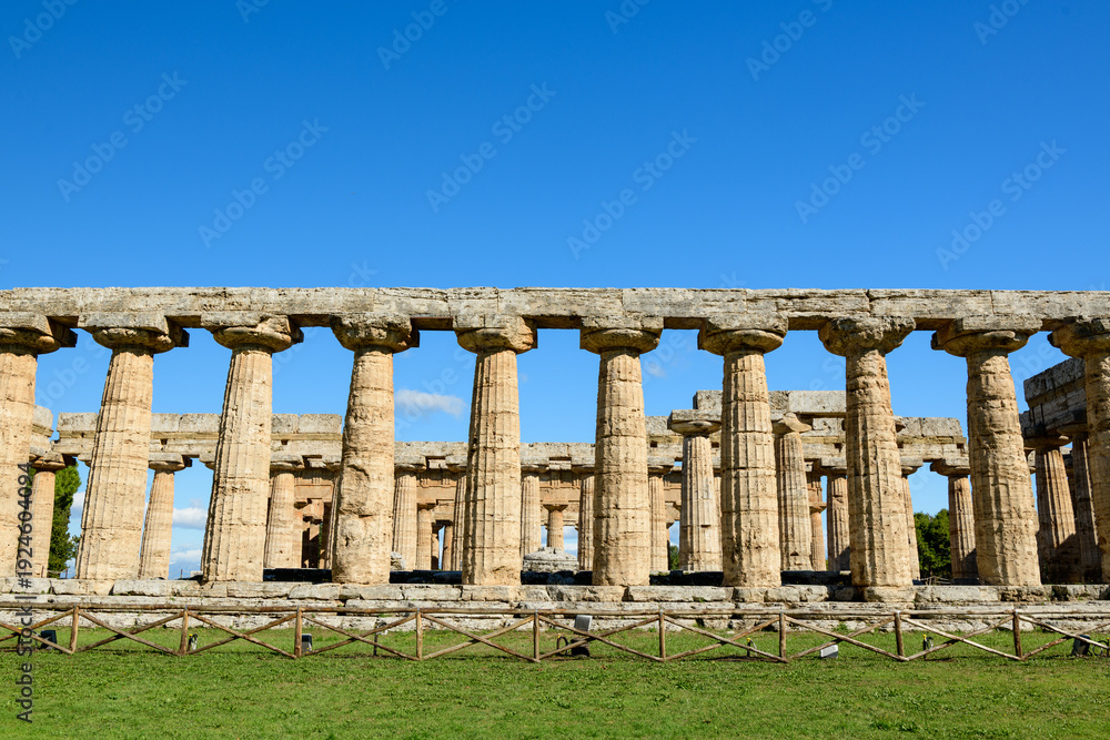 Fototapeta premium Robust Doric columns of the ancient Basilica at Paestum, Italy, form a striking linear pattern against a vivid blue sky. The weathered stone structure rises above open grassy grounds, highlighting