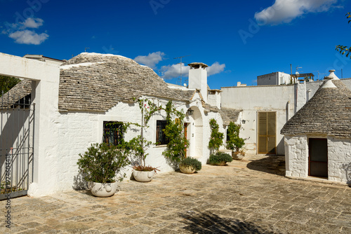 A bright stone courtyard is bordered by traditional whitewashed trullo houses with textured stone roofs in Alberobello, Apulia, Italy. Mediterranean potted plants and crisp blue sky highlight the