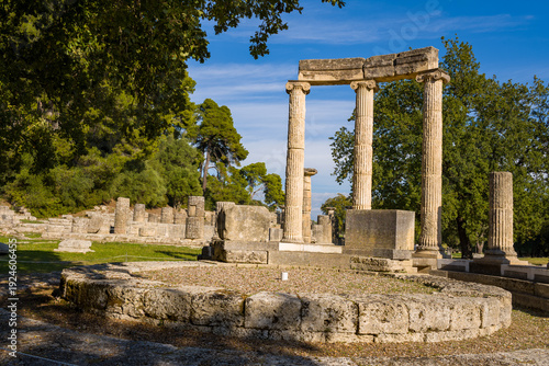 Ancient stone columns and circular foundation of the Philippeion stand amid grassy terrain and lush trees in Olympia, Greece. Afternoon sunlight highlights the classical architecture and textured