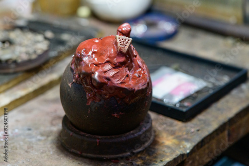 Tableau sur toile A close-up of a red wax model for jewelry making, fixed atop a dark spherical base on a worn wooden workbench in Battambang, Cambodia