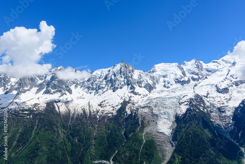 Expansive view of the Mont Blanc massif with dramatic snow-covered ridges, a sprawling glacier, and lush green forests below under a vivid blue sky with scattered clouds.