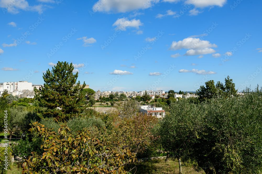 Fototapeta premium Wide view of Alberobello, Apulia, Italy, with trullo rooftops visible beyond lush trees and gardens under a bright blue sky. The scene blends Mediterranean greenery, scattered clouds, and iconic