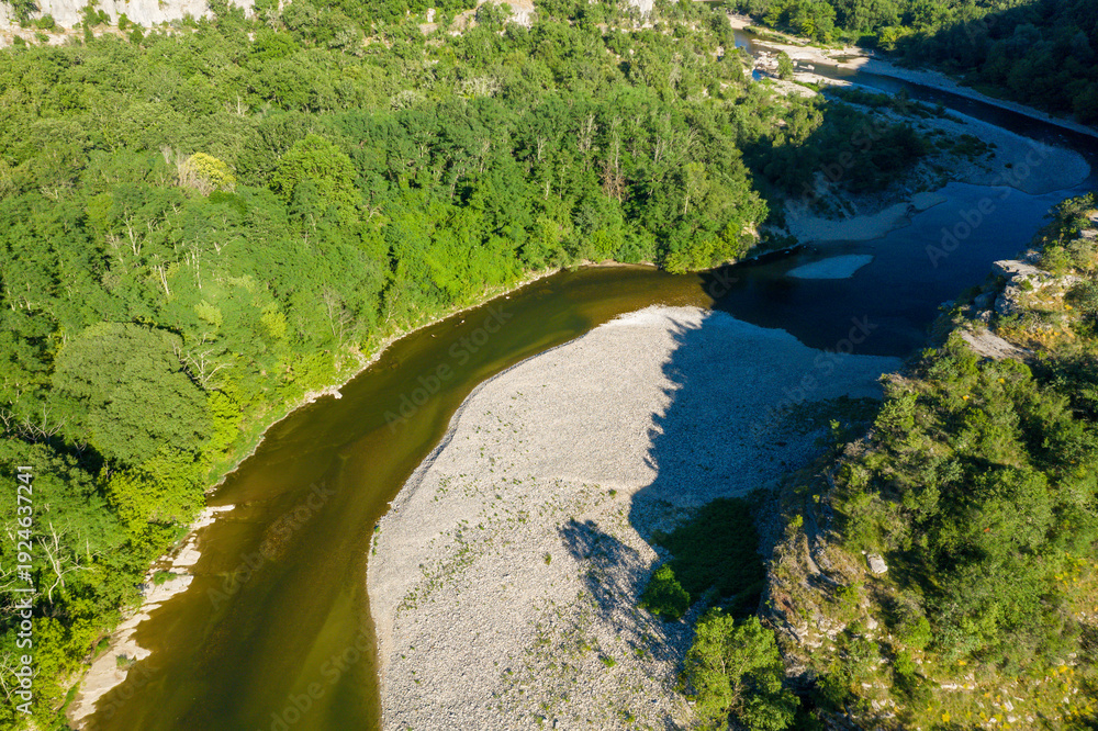 Obraz premium A winding river flows around a wide gravel bar bordered by dense green forest in the Gorges de l'Ardeche. Sunlight casts sharp shadows, highlighting the textures of rocks and foliage.
