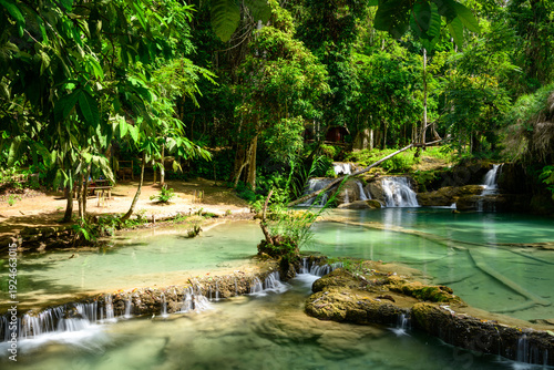 Clear turquoise water cascades over limestone terraces surrounded by dense tropical forest at Kuang Si Falls in northern Laos. Sunlight filters through lush green foliage, highlighting the natural