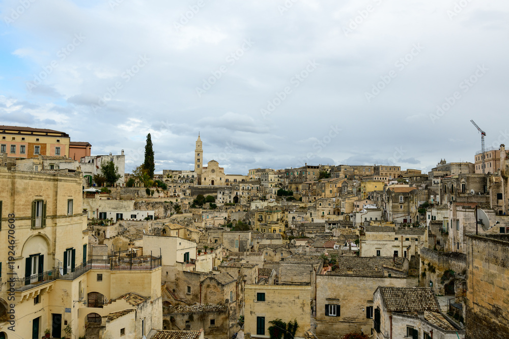 Fototapeta premium Wide view of Matera, Italy, featuring a dense array of historic stone buildings, textured facades, and the prominent cathedral tower rising above the ancient town under an overcast sky.