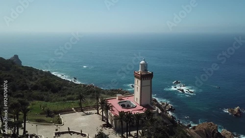 Cinematic drone shot featuring the detailed architecture of Cap Spartel lighthouse. Captures the lighthouse tower, palm trees, and the rugged coastline meeting the deep blue Atlantic Ocean.