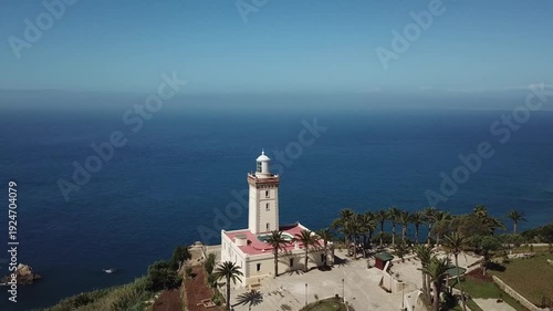 Majestic drone shot of the historic Cap Spartel lighthouse. Highlights the traditional architecture, palm trees, and the vast blue Atlantic horizon at the entrance of the Strait of Gibraltar.