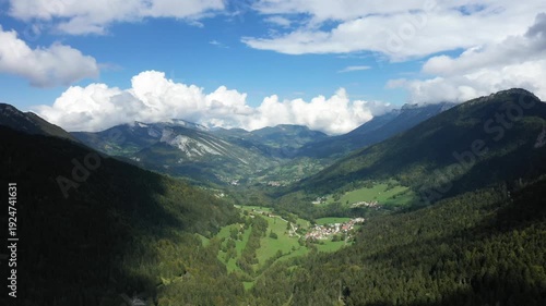 A panoramic view of a lush green valley surrounded by forested mountains in the Massif de la Chartreuse, under a partly cloudy sky. Picturesque rural villages nestle in the landscape, creating a