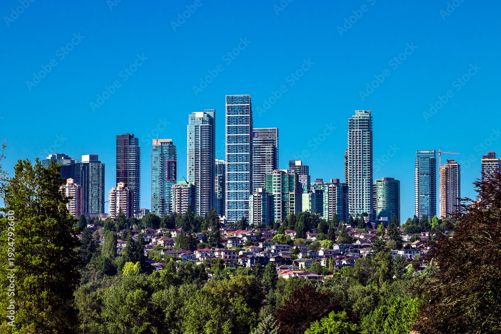 Obraz premium A residential district in Burnaby, with high rise buildings on top of a hill and private homes below, surrounded by green trees against a blue sky