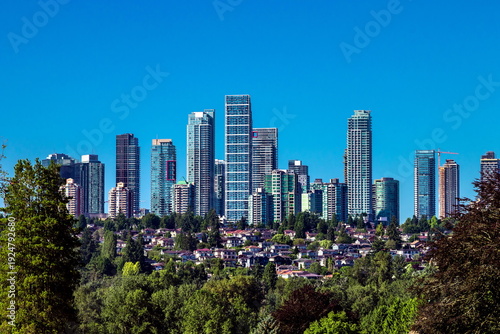 A residential district in Burnaby, with high rise buildings on top of a hill and private homes below, surrounded by green trees against a blue sky