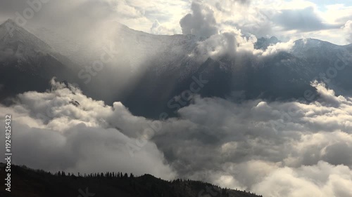Alps mountain peaks rise above cloud sea as sunrays pierce mist, cinematic landscape view. Swiss range summits overlook overcast layer, god rays through vapor, breathtaking highland vista.