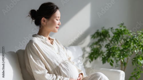 Peaceful pregnant Asian woman sitting on a sofa and meditating in a bright sunlit room.