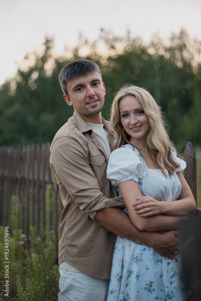 Obraz premium A young Caucasian couple stands together in a field. The man has short gray hair and wears a beige shirt. The woman has long blonde hair and a floral dress.