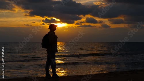 Silhouette of traveler enjoying the sunset on the beach. Solitary traveler watching a dramatic golden sunset over the ocean. The man stands on the sandy beach, observing the sun e