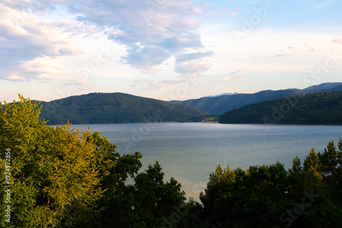 Tableau sur toile A peaceful mountain scene featuring a calm lake surrounded by lush greenery and towering hills, illuminated by soft light during the golden hour