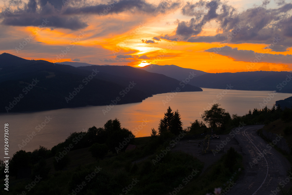 Obraz premium The sun sets behind the mountains, casting golden hues over the calm lake. Trees line the shore, creating a peaceful and picturesque mountain scene.